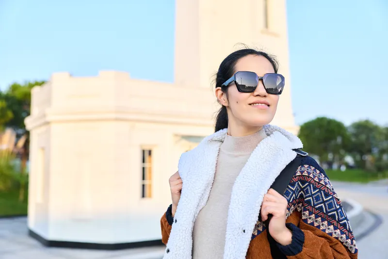 Woman wearing oversized sunglasses, scarf and layered jacket posing outdoors near a pale monument, exuding confident fashion, travel vibe and urban style