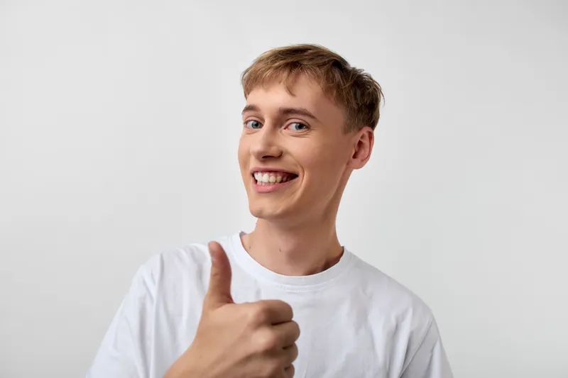 Thumbs up, smile, and casual vibe from a young man in a white t shirt in a studio setting, conveying positivity, approval, confidence, and upbeat communication for advertising and social media use
