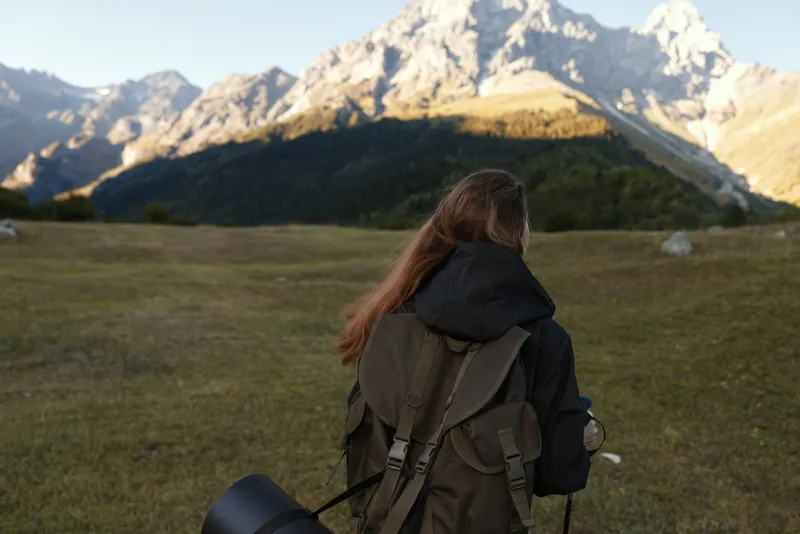 A lone photographer stands in a wide meadow with a backpack, gazing toward rugged mountains as warm sunlight blankets the scene, capturing a peaceful outdoor moment in nature.