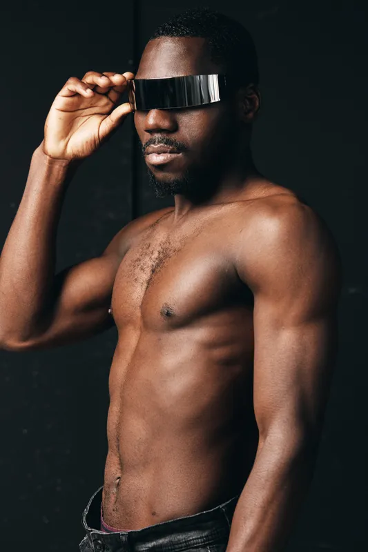 African man with muscular body and dark skin wearing futuristic visor glasses in creative conceptual visual style. Portrait of confident male model with bare chest and modern accessory.