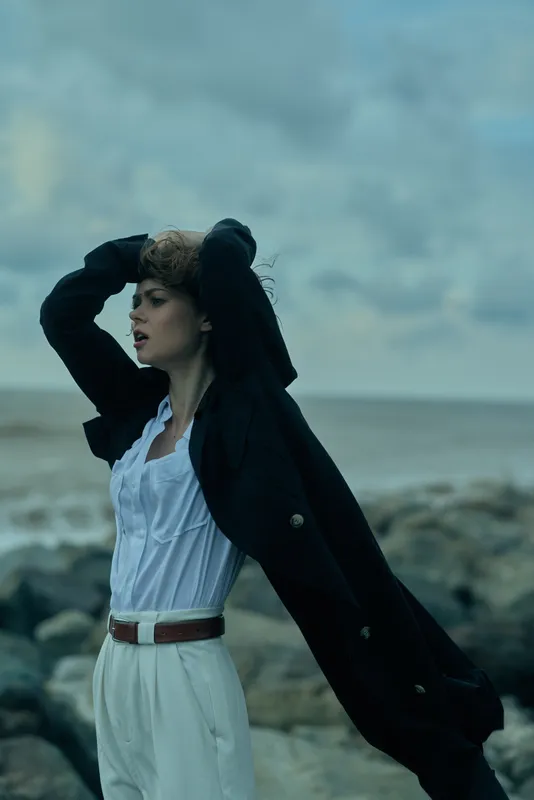 Woman in a white blouse and belt stands by the sea as a dark coat billows in the wind, creating a striking outdoor fashion moment against a rugged coast and cloudy sky