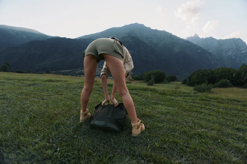 Cheerful woman outdoors bending to adjust a backpack, alpine meadow with distant mountains, warm daylight, relaxed lifestyle vibe, natural colors, fresh air, outdoor exploration