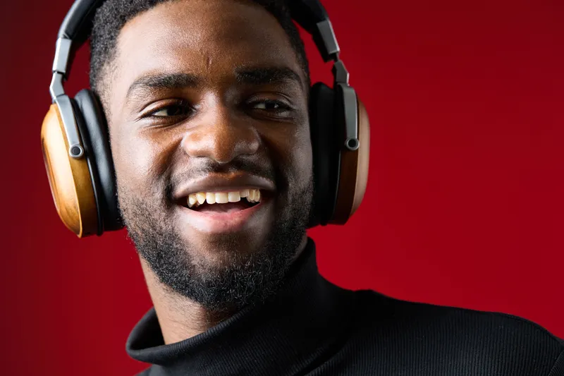 Young Black man enjoying music with headphones on, smiling, set against a vibrant red background, emphasizing joy and relaxation in a modern lifestyle