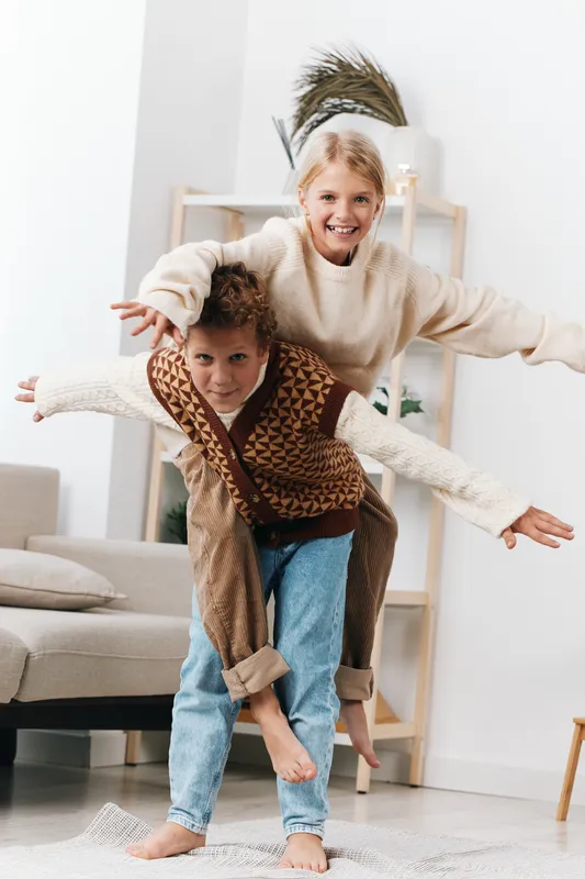 Two joyful people play indoors as a smiling woman carries a child on her back in cozy sweaters. Bright, airy living room with soft natural light highlights playful bond and carefree energy.