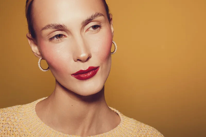 Close-up portrait of a woman with red lipstick and hoop earrings wearing a yellow sweater on a warm yellow background. Concept of beauty, fashion, and elegance.