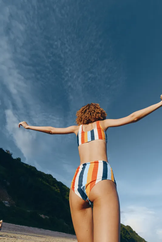 Woman, swimsuit, beach, summer, arms outstretched, standing on a coastal path, back view, blue sky and clouds, concept of freedom and vacation, natural outdoor light