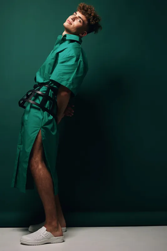 Young man in futuristic corset belt and green dress with short sleeves standing against emerald backdrop, posing sideways with serious face and white slip-on shoes on clean background.