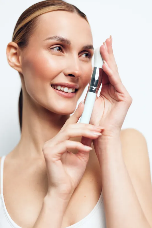 Young smiling woman with clean skin holding white cosmetic brush near face applying makeup, isolated on white background, beauty skincare concept with natural light and fresh look.