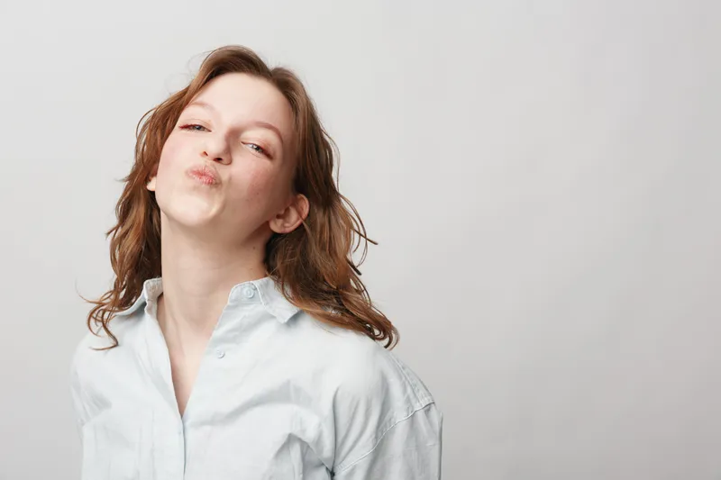 Young woman with red hair wearing white shirt blows a kiss with puckered lips. Studio portrait on light background. Concept of affection, flirtation, and playful mood.