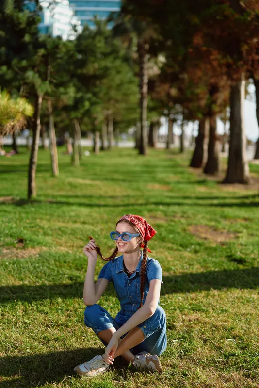 Lifestyle woman in modern boho-western denim street style sitting in park with sunglasses and red bandana, captured in warm film color lighting outdoors.