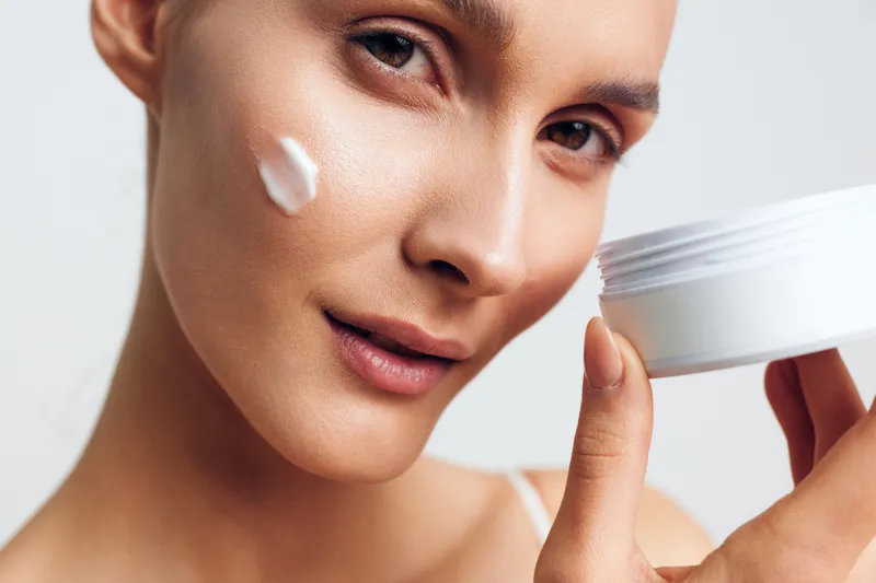 Young woman with clear skin applies white face cream on her cheek while holding an open jar, expressing gentle care and a calm mood in a bright studio setting.