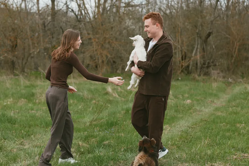 A young man holds a white baby goat while a young woman reaches out to pet it in a grassy field with bare trees, capturing a candid moment of connection and lifestyle outdoors.