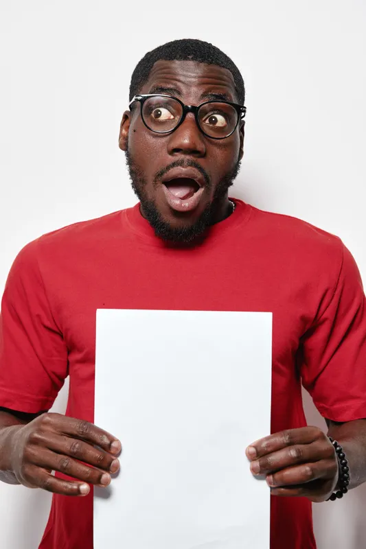 African American man with dark skin and glasses wearing red shirt holding blank white paper with surprised expression on white background studio portrait.