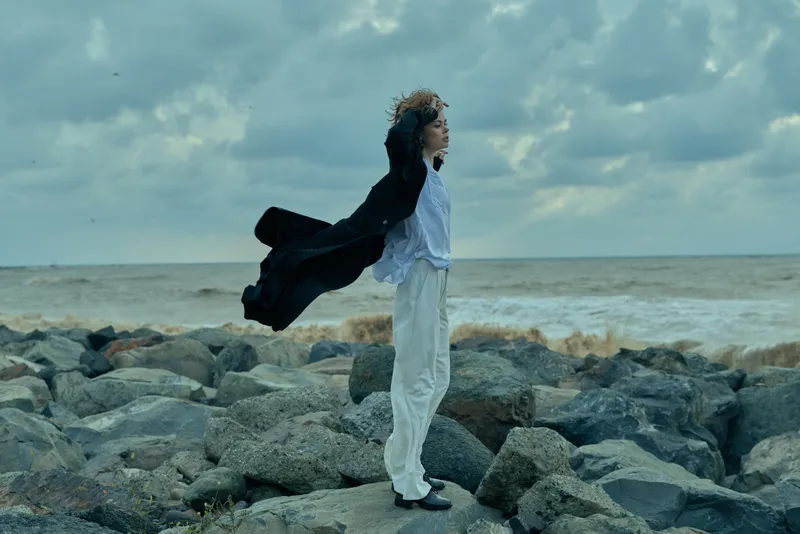 Wind, ocean, coast, rocks, fashion, cape, outdoor, model, windblown, dramatic scene shows a figure on a windswept shoreline, cape billowing, light apparel, motion and resilience against a cloudy