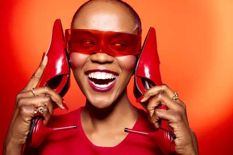 Happy African woman with dark skin wearing red dress and sunglasses holds red high heels near face on orange background. Concept of fashion, style, beauty, and joyful emotions.