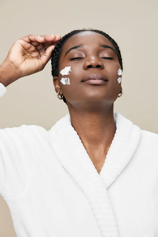 Woman applying skincare cream, showcasing a natural beauty routine with closed eyes, warm skin tone, and a serene expression against a light beige background