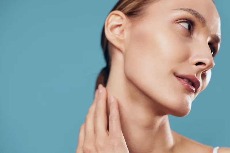 Close-up of a young woman with clear skin touching her neck against a blue background. Concept of skincare, beauty, wellness, and natural self-care with soft lighting.