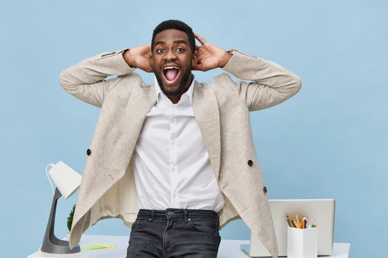 Joyful young African American man expressing excitement and surprise while standing at office desk, hands on head, smiling, wearing casual beige blazer and white shirt, with colorful stationery and la