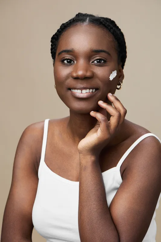 Smiling Black woman with skincare cream on her face, wearing a white top, showcasing healthy skin and beauty in a minimalist beige background