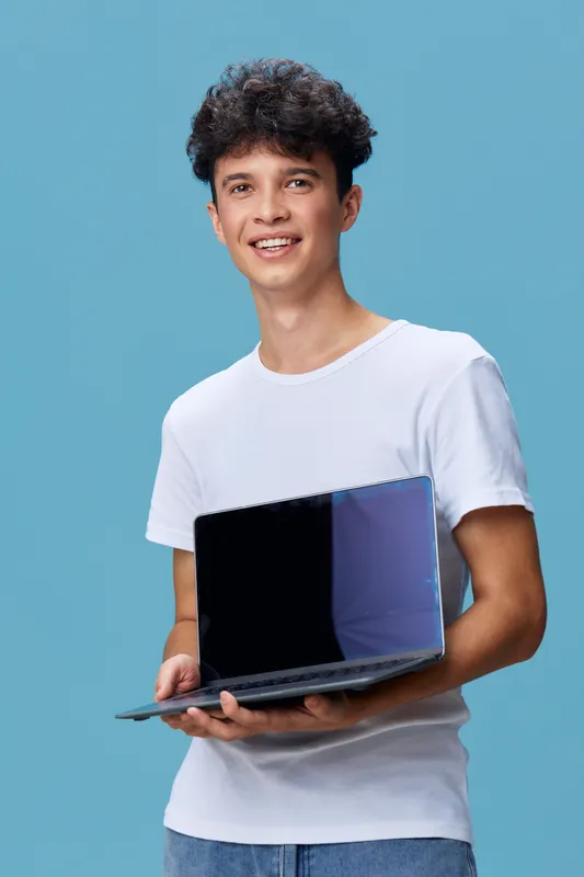 smiling man holding laptop, casual young adult with curly hair wearing white t-shirt on blue background, expressing happiness and confidence.