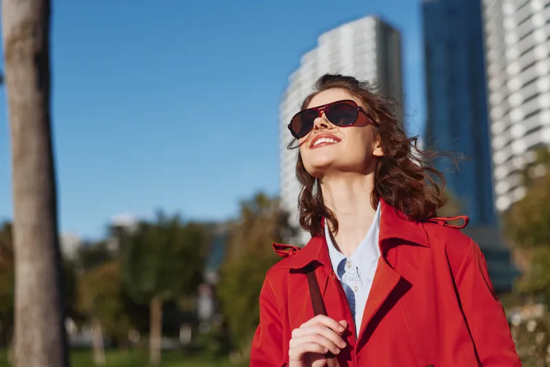 Young woman in a red coat smiles at the sun in a bright urban park, wearing sunglasses and enjoying warm weather, with modern buildings rising behind her.