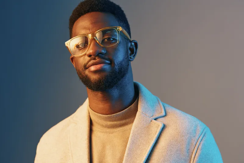 Stylish portrait of a young African man wearing glasses and a beige coat, exuding confidence and sophistication against a modern blue background