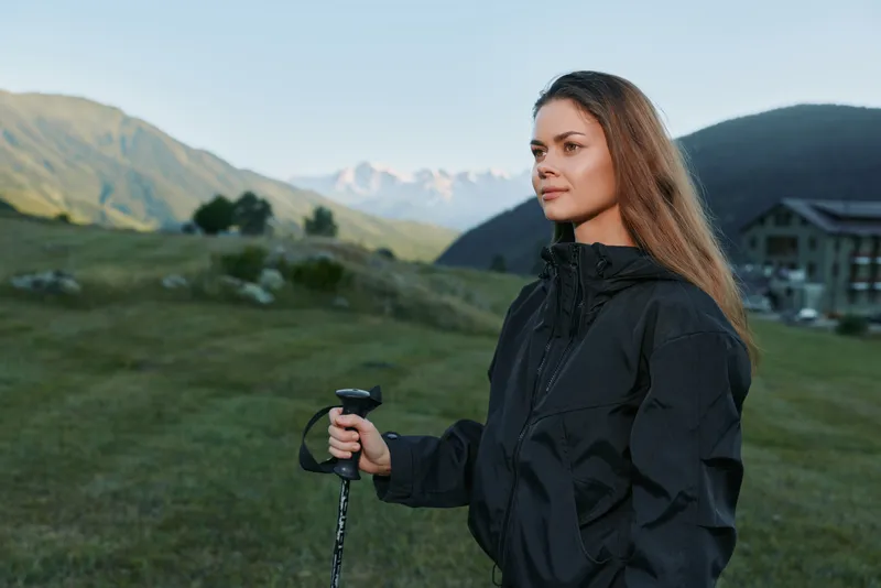 A contemplative woman stands in a alpine meadow with a trekking pole, wearing a dark rain jacket. She gazes toward distant peaks, conveying calm adventure and quiet resilience.
