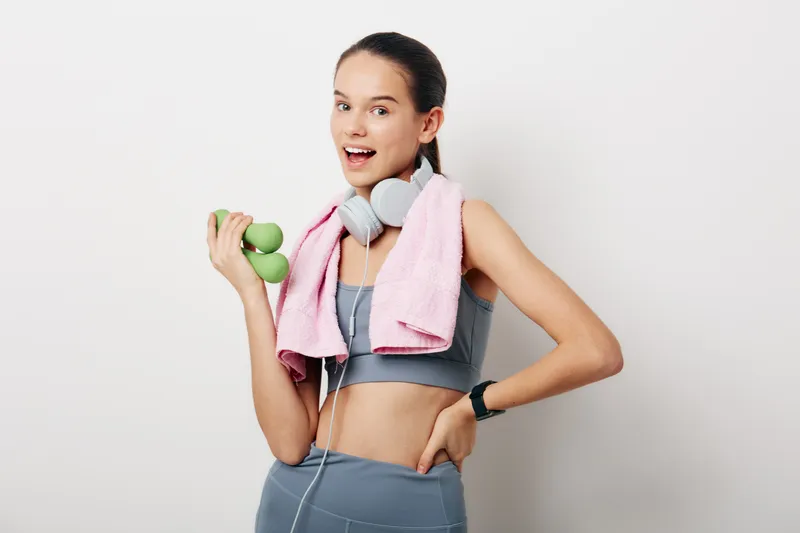 Athletic woman in gym wear lifts green dumbbells, with a pink towel over shoulders and headphones, showcasing fitness, sport and workout energy in a bright studio setting.