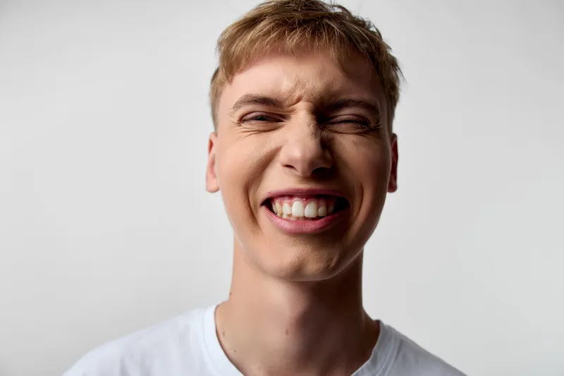 Close up of a young man with a wide grimace showing teeth, conveying intense emotion and awkward energy, studio light, plain background, raw facial expression highlighting mood and spontaneity