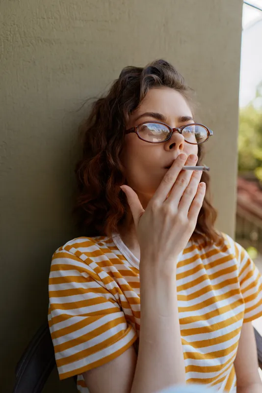Cheerful woman outdoors, striped shirt, glasses, smoking, relaxed pose by a wall, warm sunlight, natural colors, casual lifestyle moment, confident and thoughtful in everyday setting
