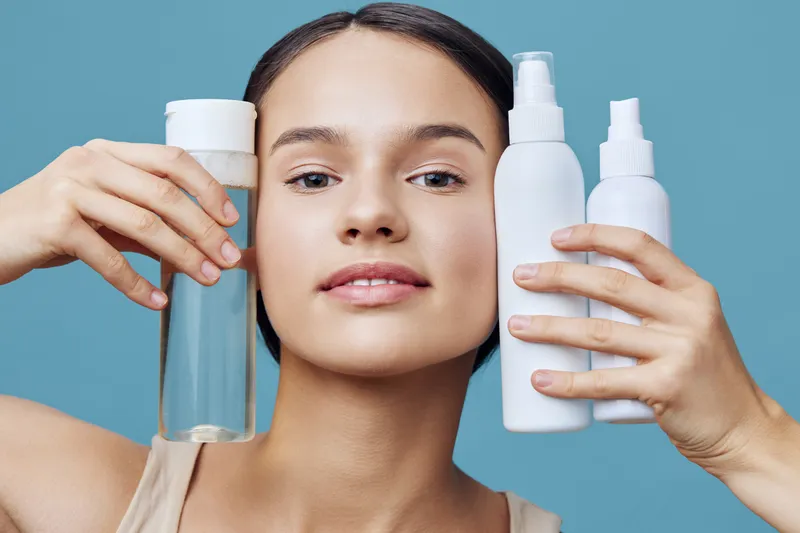 Young woman showcasing skincare products with a serene expression against a soft blue background, promoting healthy skin and beauty rituals