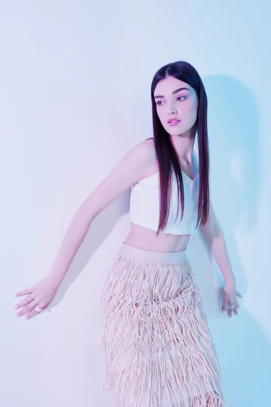 Young woman with long straight hair poses in a white crop top and fringed skirt against a pastel background. Concept of beauty fashion and modern style.