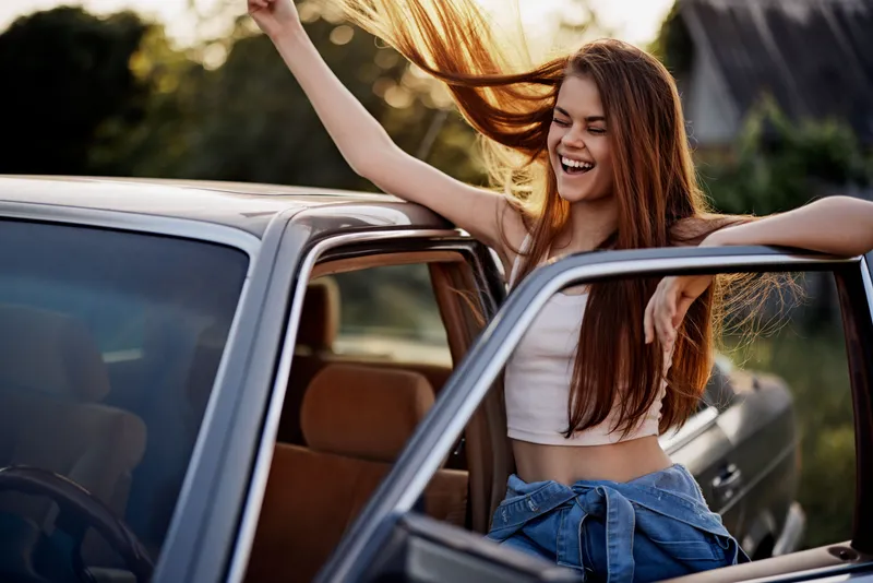 Joyful young woman excitedly enjoying a sunny day while leaning out of a car, showcasing her long brown hair and casual summer outfit.