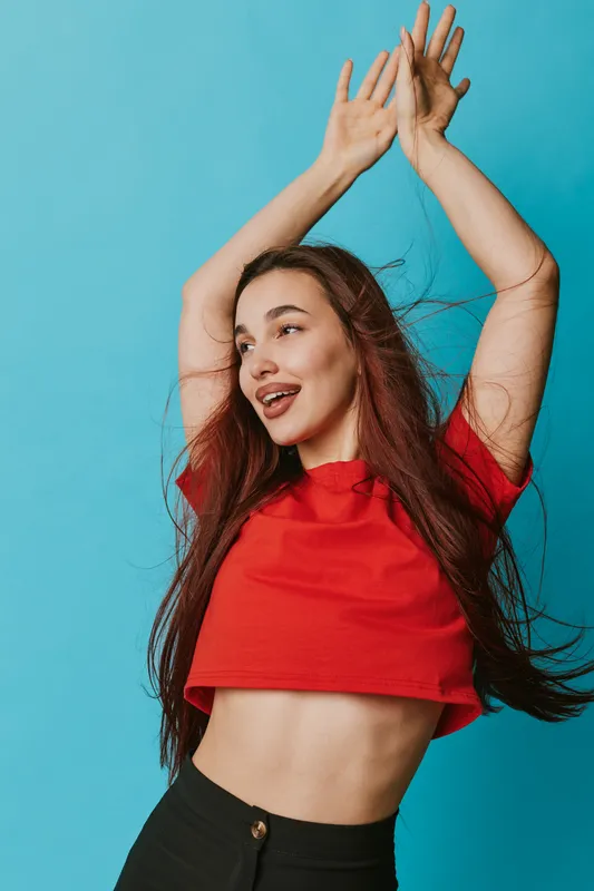 Young woman in red crop top dancing with raised arms and long hair flowing against blue background. Casual style, joyful expression and movement in studio portrait.