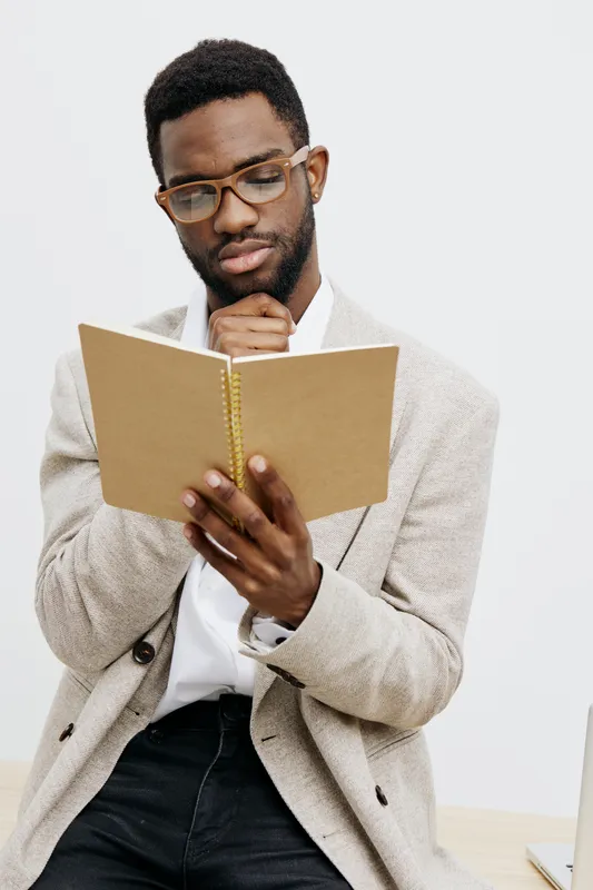Thoughtful young African American man wearing glasses and casual beige cardigan holding a notebook, looking pensive as he sits against a plain white background. People lifestyle concept, student, refl