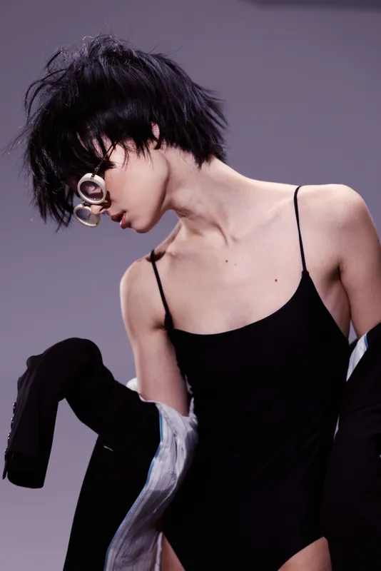 Stylish woman with short black hair and sunglasses wearing a black outfit in a minimalist studio setting.