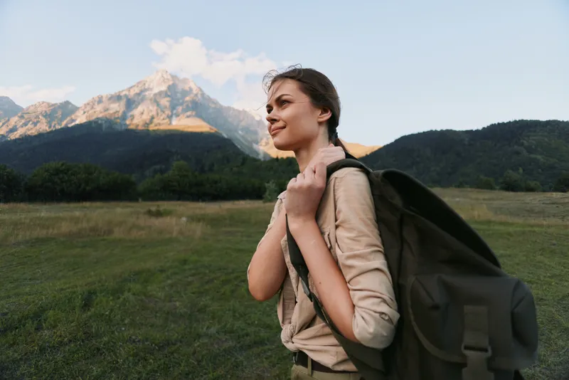 Woman with backpack hiking outdoors in a mountain meadow, exuding confidence and curiosity. Concept of travel, exploration, wellness, and nature in warm daylight, soft glow.
