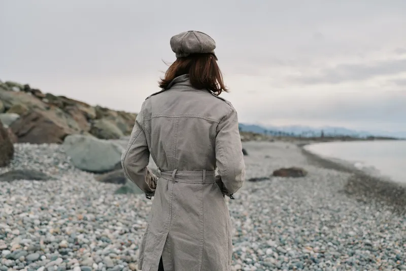 Woman in a patterned coat and beret stands on a rocky beach looking at the cloudy sky. Concept of autumn fashion, solitude, and nature exploration in cool weather.