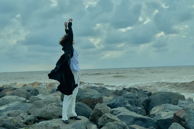 Woman stands on rocky shore with arms raised in celebration, wind whipping clothing, dramatic sea horizon and moody clouds creating an outdoor freedom moment