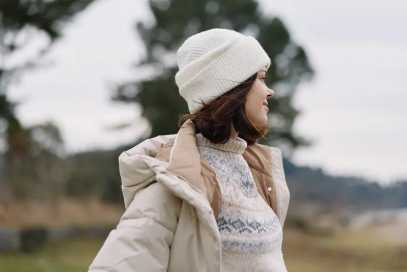 Woman in a warm beige coat and knit sweater wearing a white beanie stands outdoors in a windy open landscape, showcasing winter fashion and relaxed style against a natural backdrop