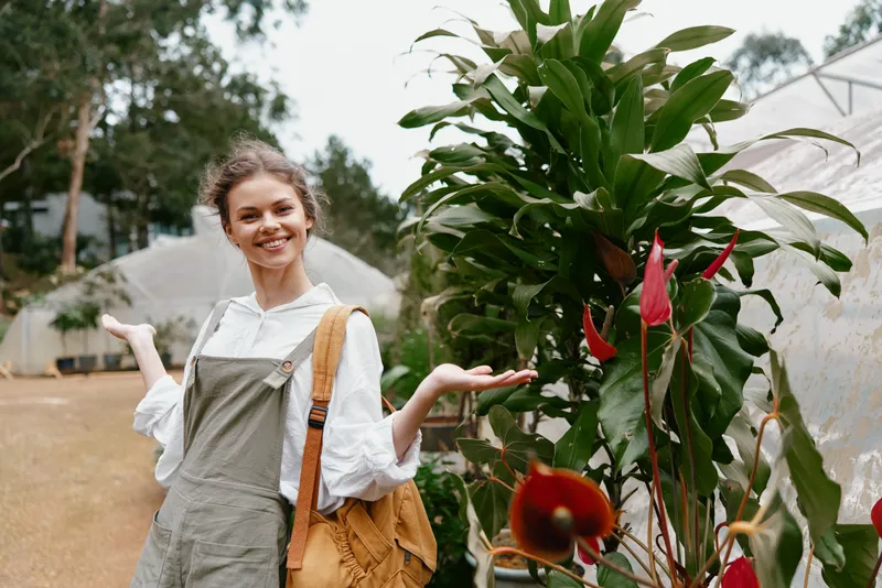 Happy woman in casual outfit showcasing greenery in a botanical garden, wearing overalls with a bright smile amidst tropical plants and flowers