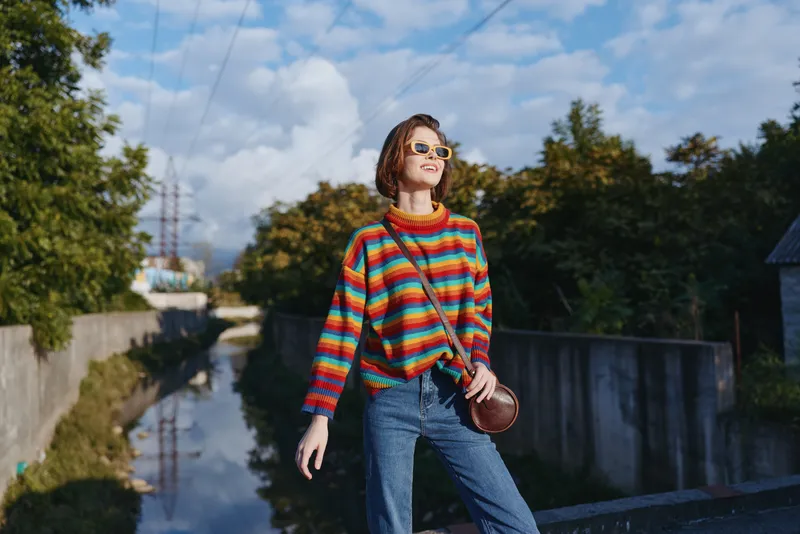 woman striped sweater sunglasses crossbody bag jeans smiling urban canal walking in casual outfit, young short brown hair millennial enjoying sunny lifestyle travel and colorful knit fashion