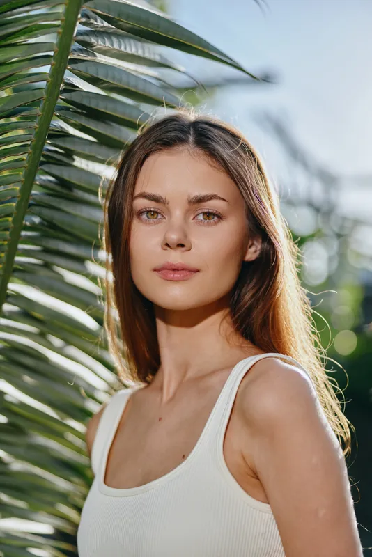 young woman portrait with long brown hair wearing white tank top standing near tropical palm leaves in natural sunlight with serene expression and blurred background