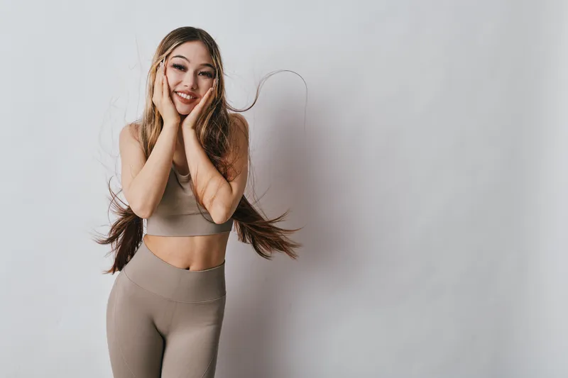 Young woman smiling in beige sportswear with long hair posing on gray background for fitness, health, wellness, and active lifestyle concept in studio.