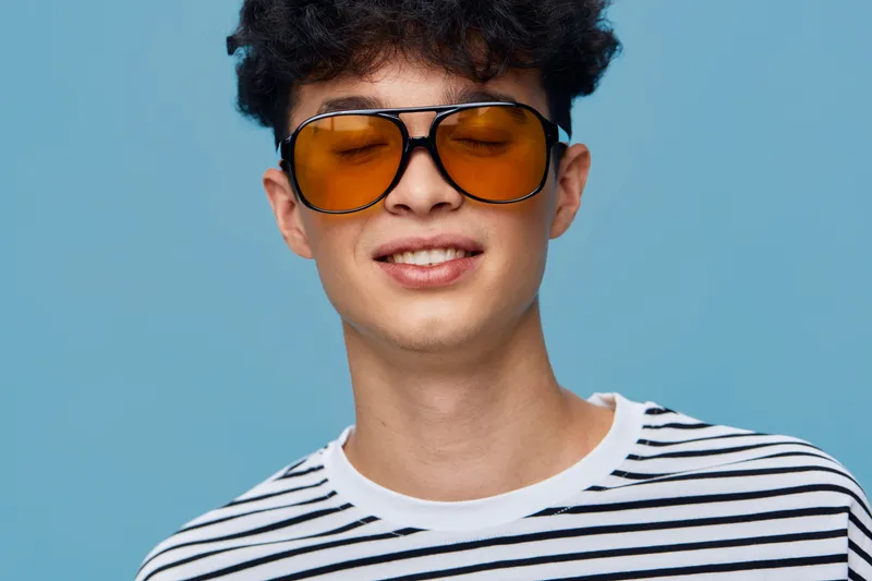 young man wearing yellow sunglasses with striped shirt smiling against blue background, casual style, modern and cheerful expression