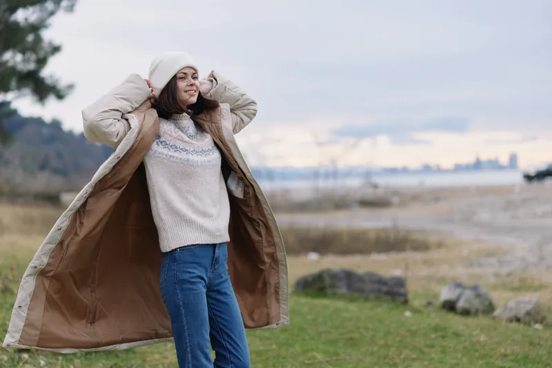 Woman outdoor coat wind landscape casual sweater autumn lifestyle, relaxed pose in a scenic natural field by coastline with breeze and peaceful daylight.