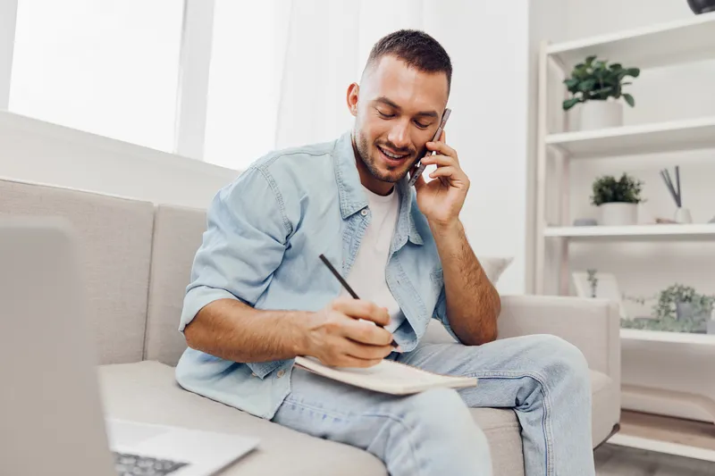 Happy man talking on the phone while taking notes in a cozy modern living room, showcasing a blend of productivity and comfort.