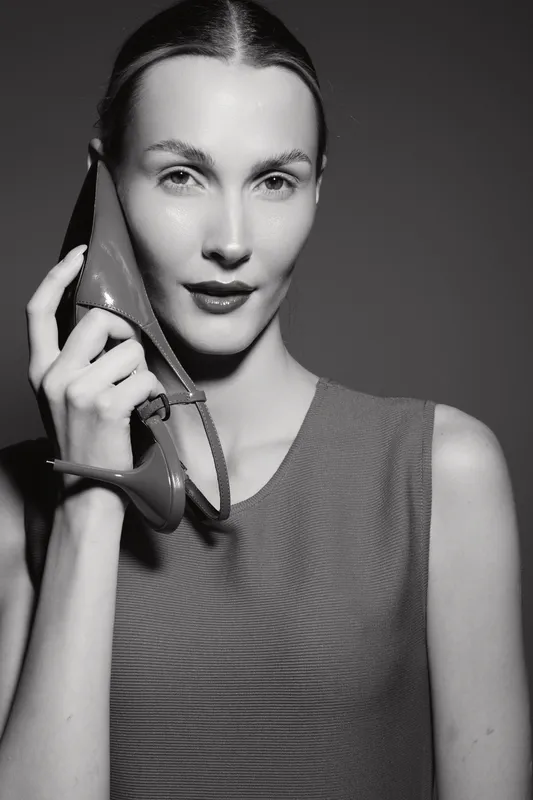 Young caucasian woman with fair skin holds high heel shoe near face in black and white studio portrait. Concept for fashion, beauty, style, and elegance.