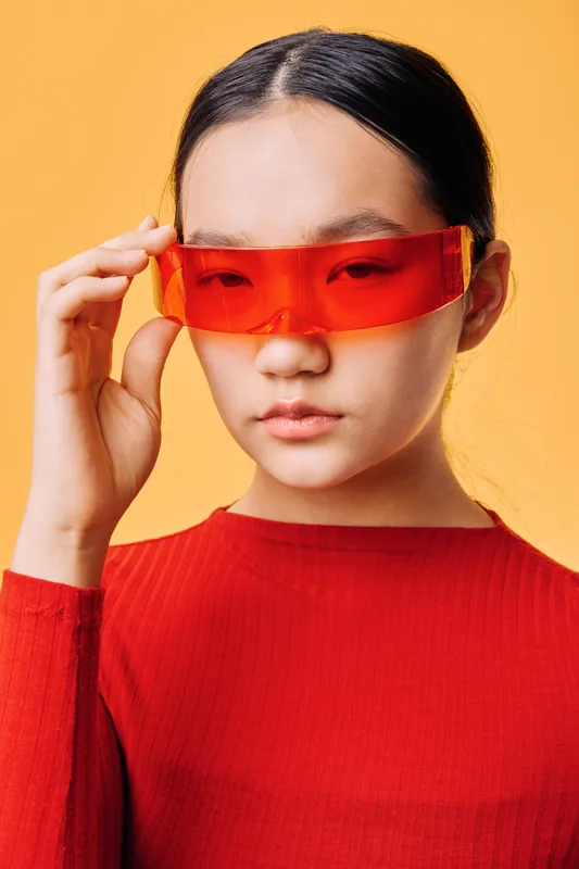 Asian woman with long black hair wearing red futuristic sunglasses and red ribbed sweater standing against yellow background in studio portrait with serious look and minimal makeup.