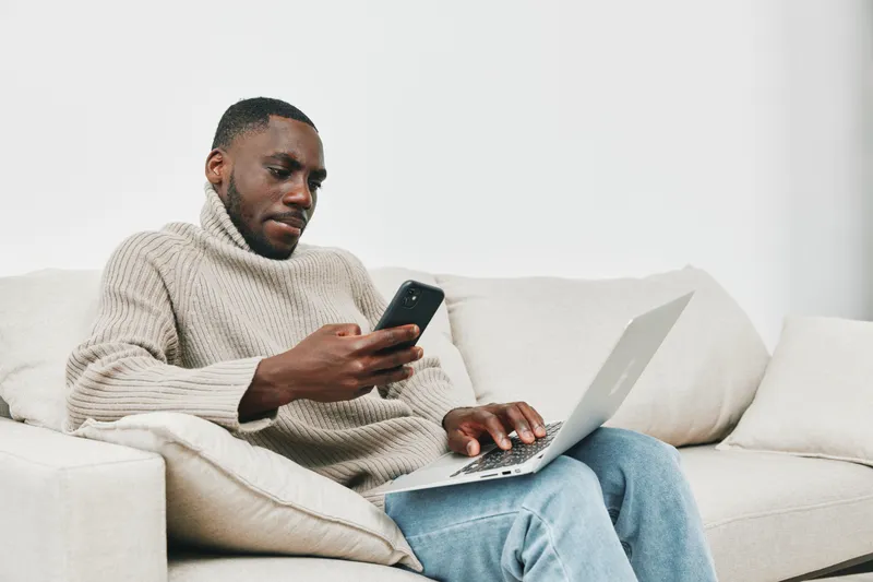 Young Black man in a cozy sweater engaging with smartphone and laptop, sitting on a beige couch in a minimalist living space